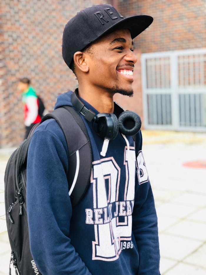 Smiling young man wearing cap and headphones, standing with backpack outdoors.