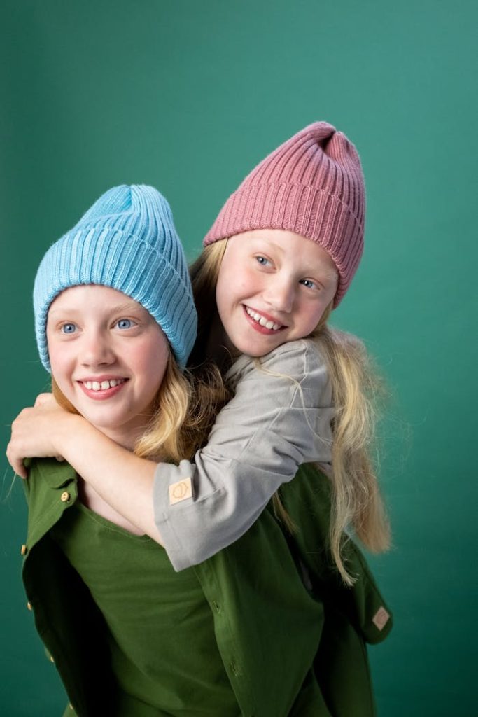 Two young girls having fun with a piggyback ride, wearing colorful hats, against a green background.